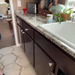 Dark brown kitchen cabinets with white knobs, granite countertop, white sink, and hexagonal tile floor.