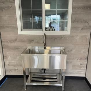 Stainless steel utility sink with tall faucet against grey wood-paneled wall under a window.